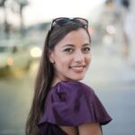 Cheerful woman in a purple top smiles at the camera on a bustling city street.